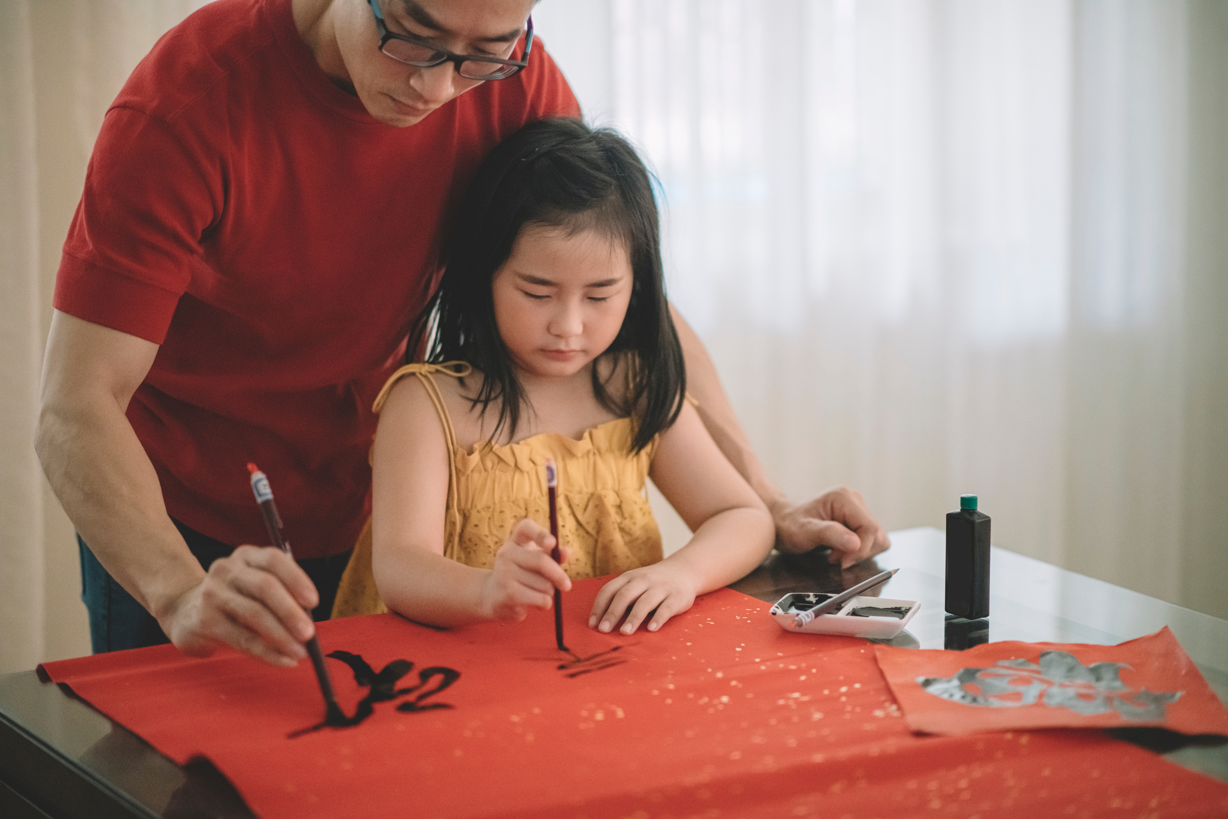 an asian chinese male practising chinese caligraphy for coming chinese new year celebration home decoration purpose with prosperity and good wording by writing it on a red piece of paper and teaching his daugther