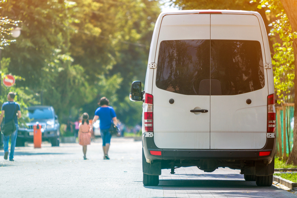 Back View of White Passenger Medium Size Commercial Luxury Minibus Van Parked N Shadow of Green Tree on Summer City Street I with Blurred Silhouettes of Pedestrians and Cars under Green Trees.